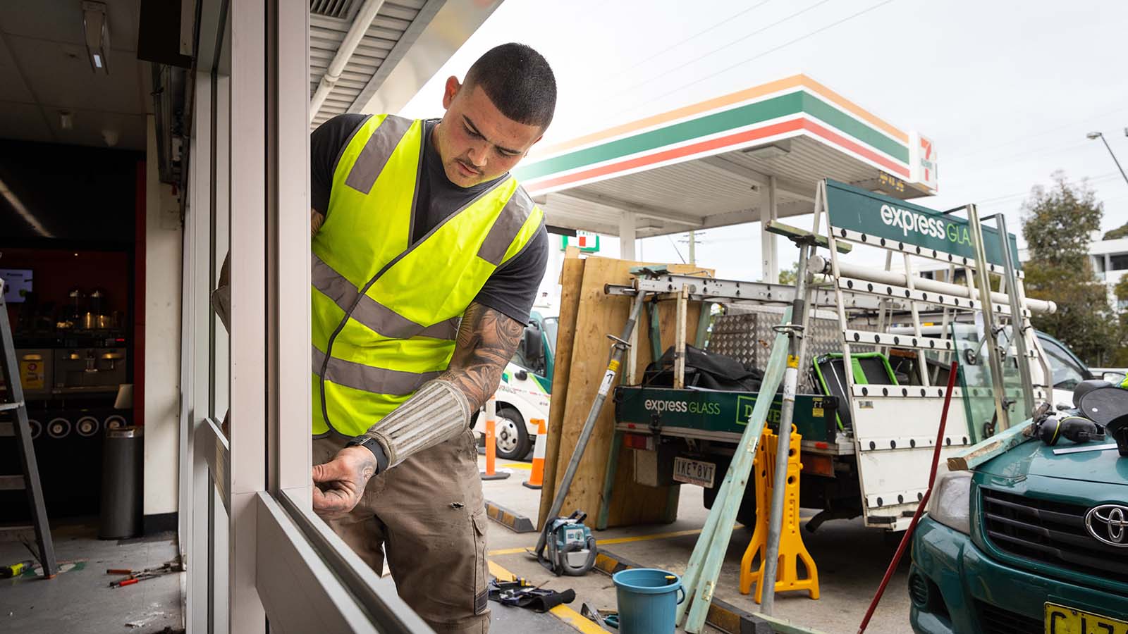 An Express Glass expert glazier in a high-visibility vest is working on a broken window at 7-Eleven Pendle Hill, using tools. In the background, there are construction materials and an Express Glass work vehicle.