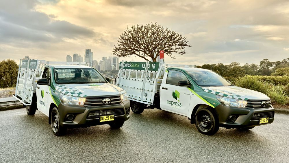 Two Express Glass service vehicles parked on a wet road, with a city skyline visible in the background. The vehicles have a tree on the back of one and are marked with the Express Glass logo and green and white designs.